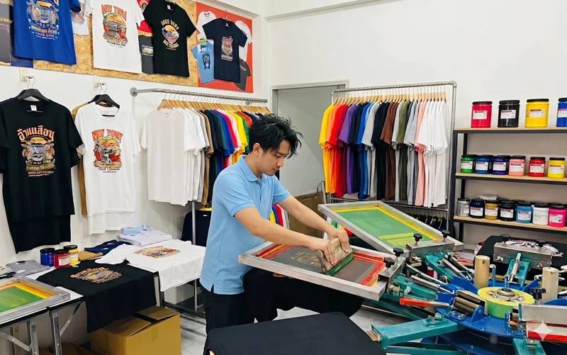 ภาพผู้ชายสวมเสื้อโปโลสีฟ้ากำลังทำการสกรีนเสื้อ ลายต่างๆลงบนเสื้อยืดด้วยเครื่องสกรีนแบบมือหมุน ภายในร้านสกรีนเสื้อที่มีเสื้อยืดสกรีนลายแขวนโชว์บนผนังและมีราวแขวนเสื้อยืดเปล่าหลากสีอยู่ด้านหลัง รวมถึงชั้นวางกระปุกสีสกรีนจำนวนมาก