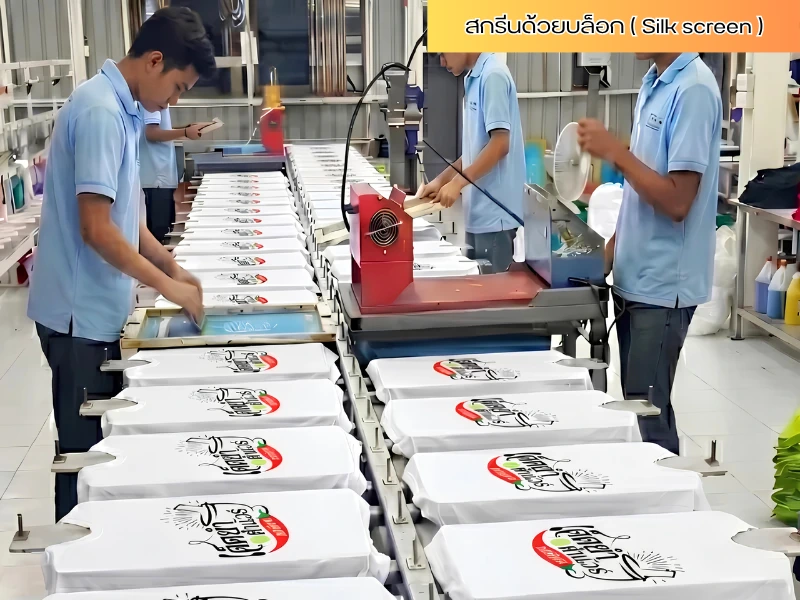ช่างพิมพ์กำลังใช้บล็อกสกรีนสกรีนลายลงบนเสื้อยืดสีขาวบนโต๊ะพิมพ์สกรีนยาวระบบ Silk Screen ในโรงงาน